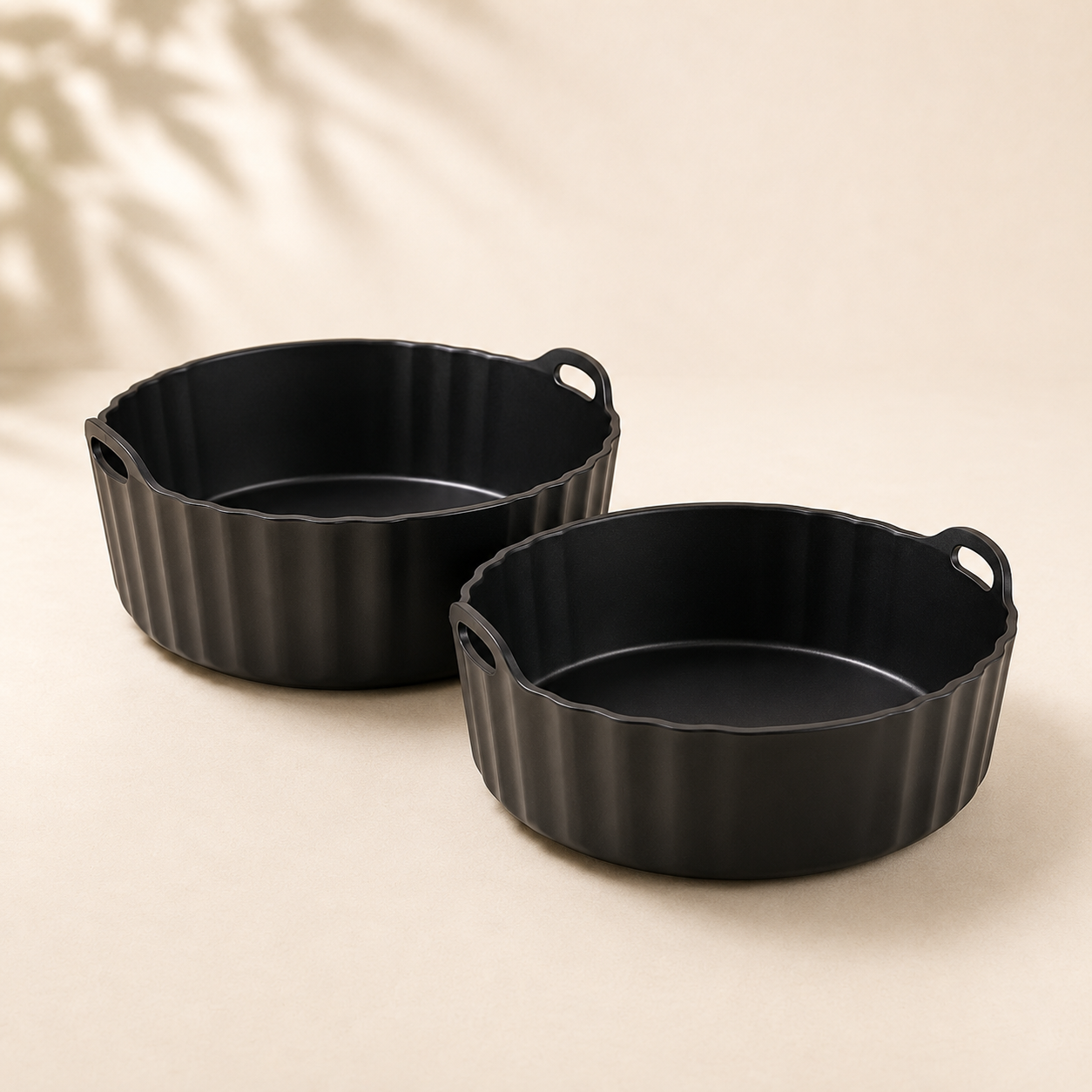 Zwei schwarze runde Airfryer-Backformen mit Griffen vor hellem Hintergrund